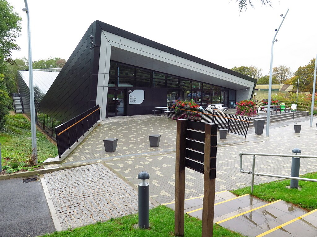 Modern building with angular design and outdoor seating area, surrounded by greenery. | Battle of Britain Bunker Exhibition Centre, Uxbridge by Andrew Curtis, CC BY-SA 2.0, via Wikimedia Commons