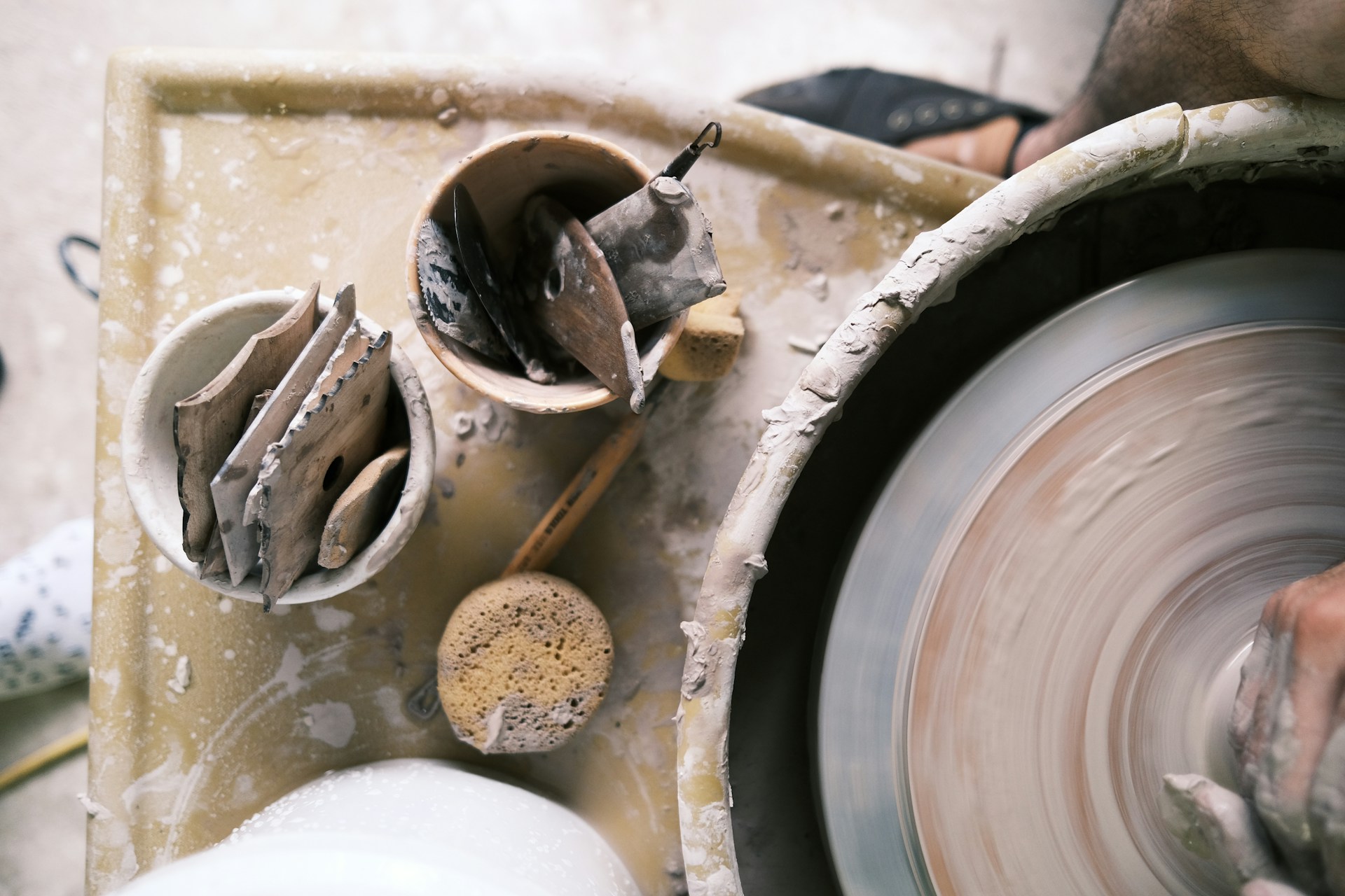 Pottery tools on a messy table beside a spinning potter's wheel with a person's hands working. | Photo by Taylor Heery on Unsplash