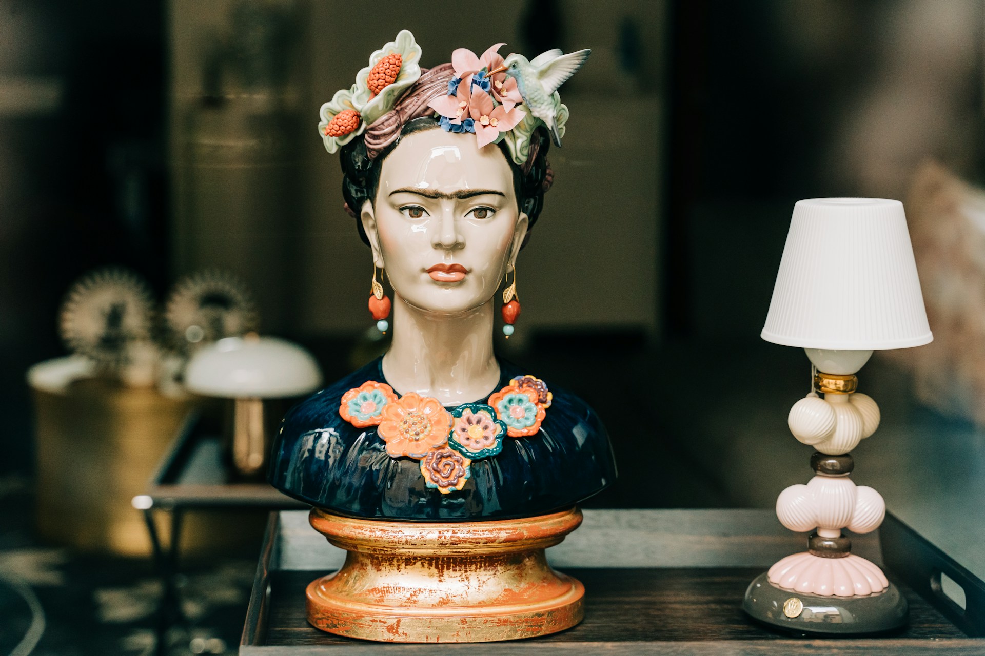 A decorative bust with colorful flowers and a bird on its head next to a table lamp. | Photo by Gabriella Clare Marino on Unsplash