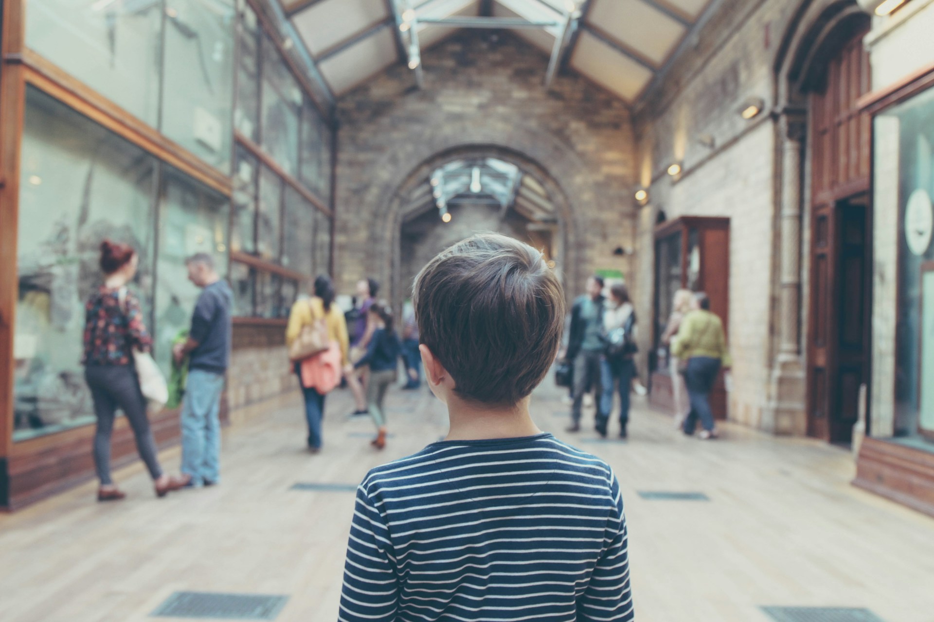 Child in striped shirt observing people in a gallery with exhibits on display. | Photo by Michał Parzuchowski on Unsplash