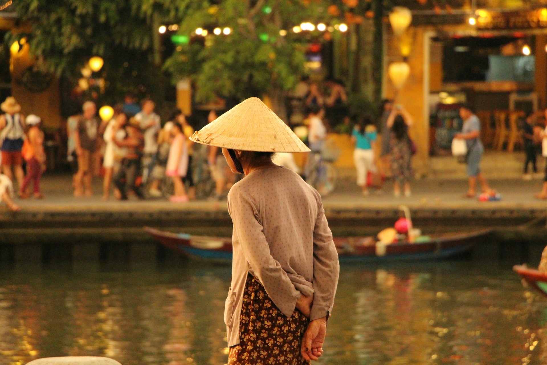 Person in conical hat facing a bustling riverside crowd in the evening. | Photo by Katherine McCormack on Unsplash