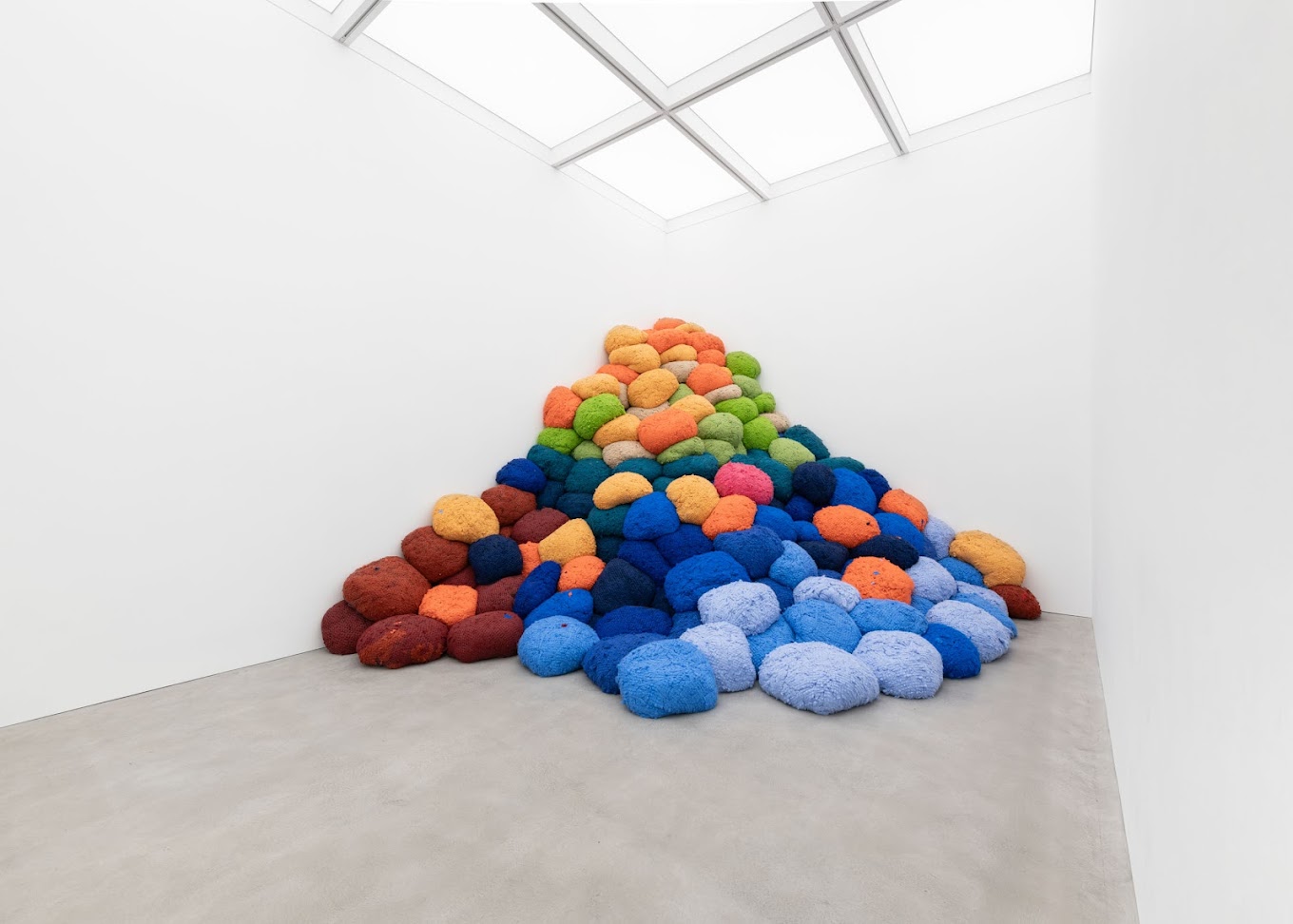 Colorful spherical objects arranged in a pyramid shape in a white gallery room with a skylight. | Alison Jacques 2024