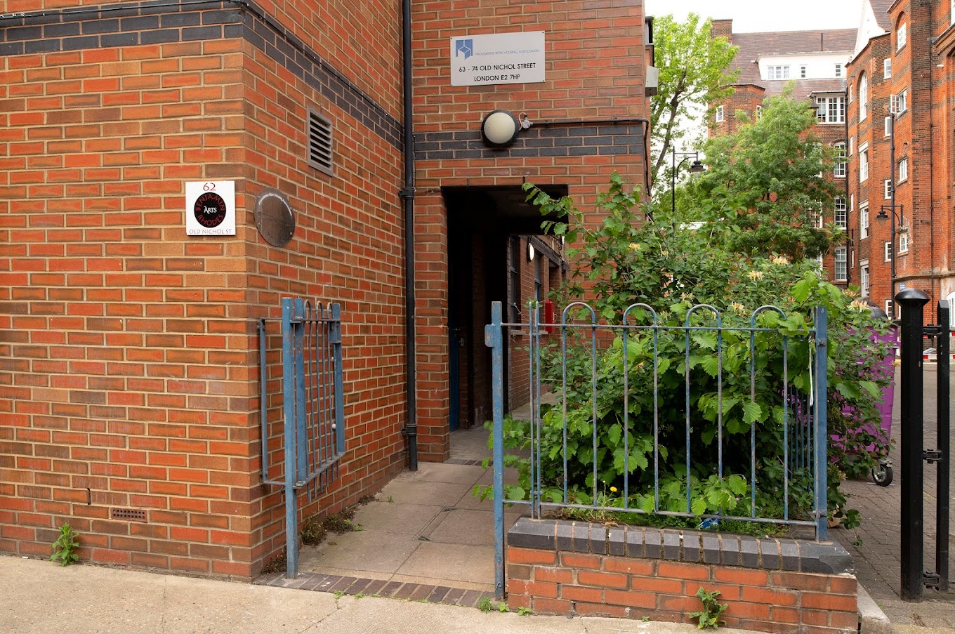 Red brick building with a gate outside and some plants. | Benjamin Rhodes Arts