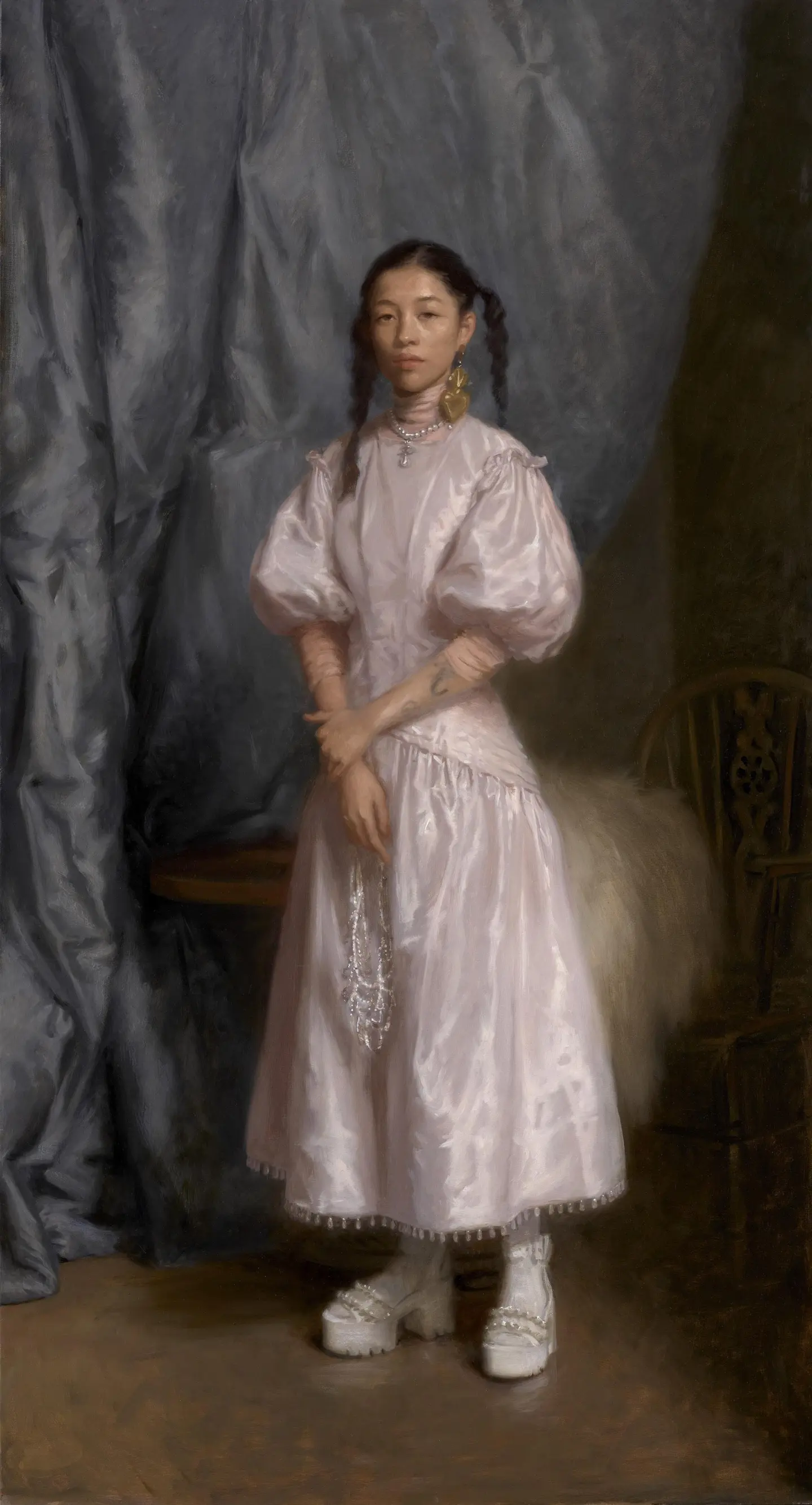 Portrait of a girl in a pink dress, standing with hands clasped, against a draped background.