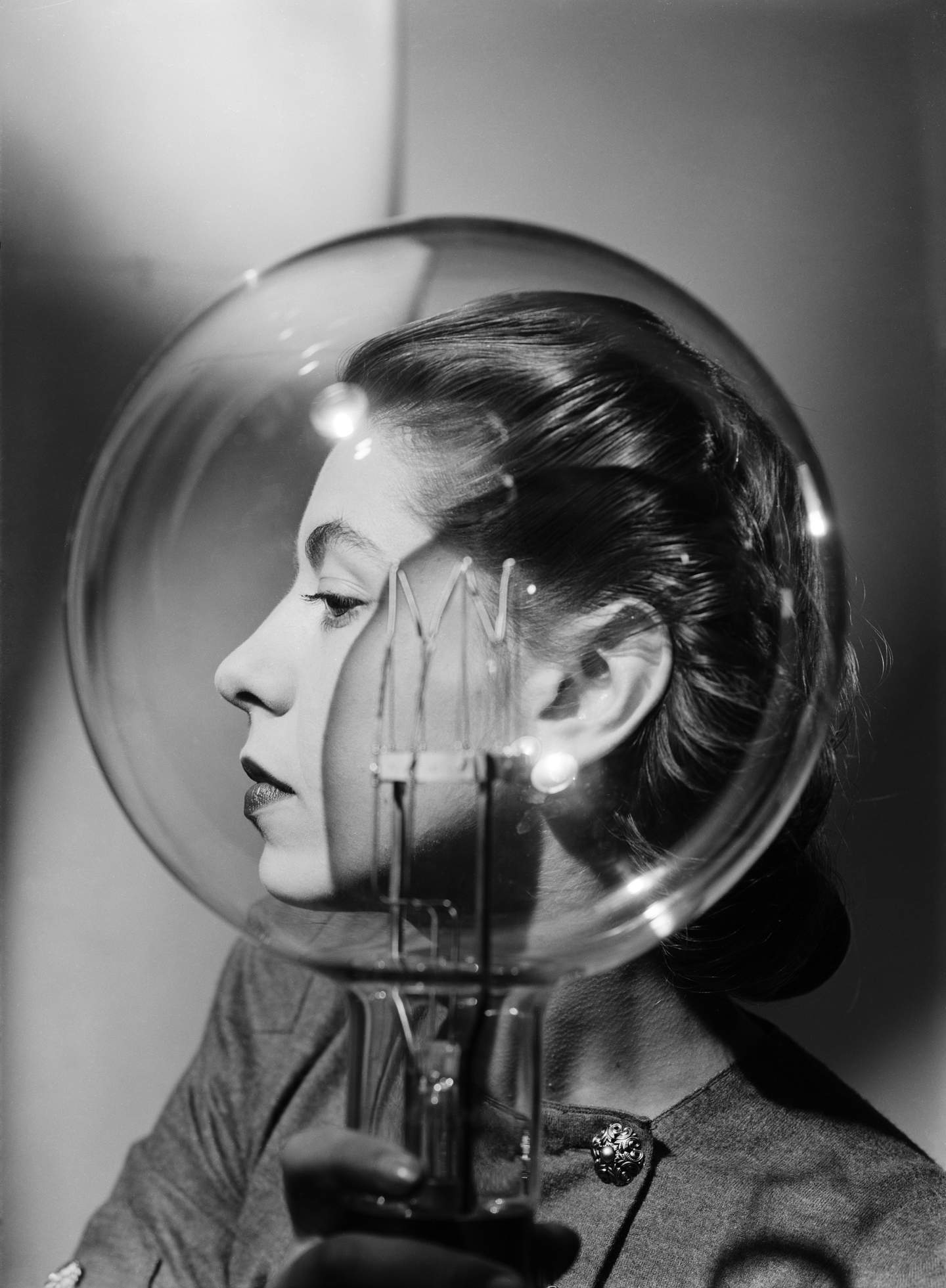 Person holding a large transparent bulb in front of their head, wearing a pearl earring and brooch.