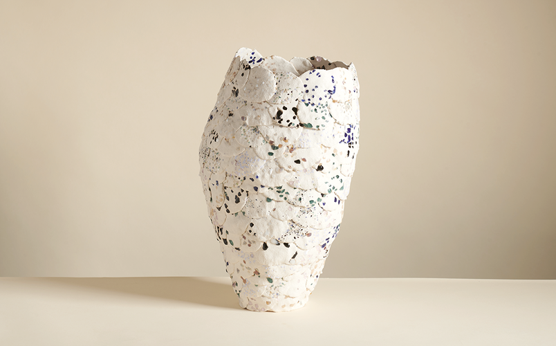 An asymmetrical, textured vase with speckles of color on a beige background.