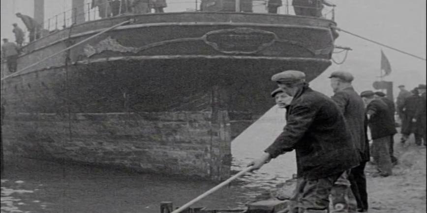 A black and white photo of a vintage ship docked with people standing nearby.