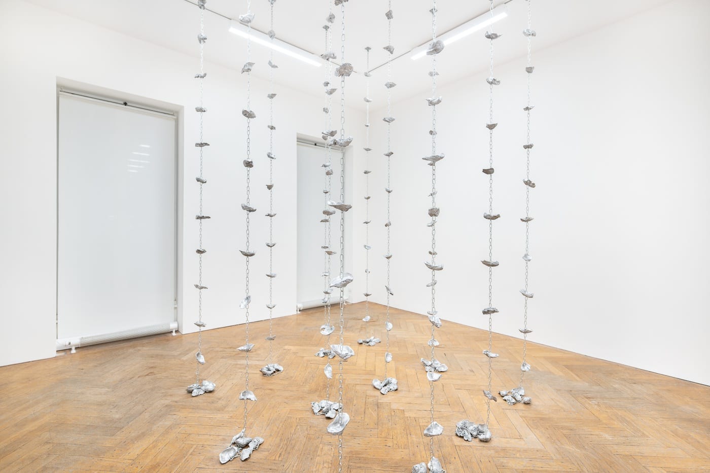Modern art installation with hanging chains and attached rocks in a white gallery space with wooden floors. | 2024 Ben Hunter