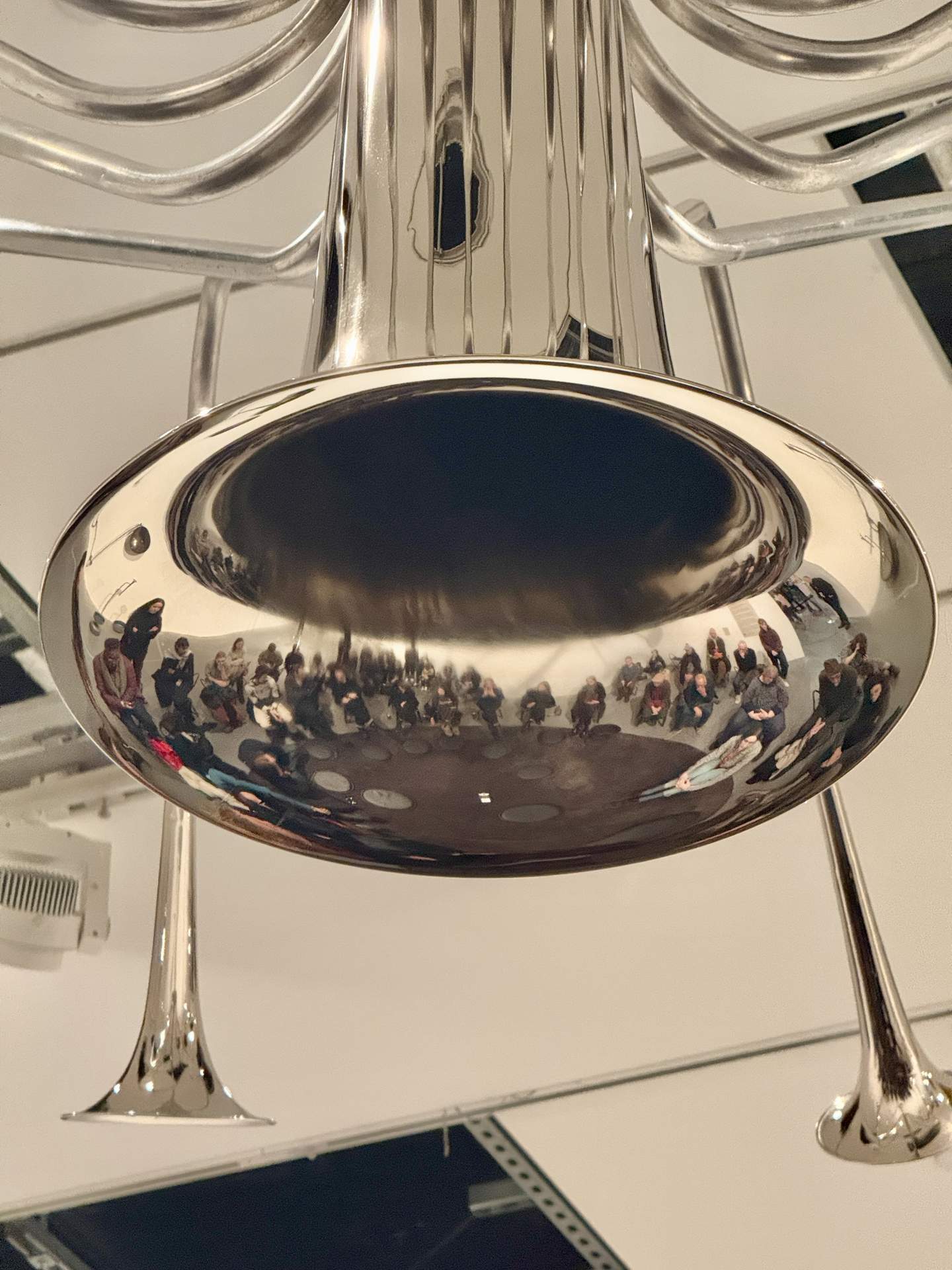 A reflective metallic instrument captures the images of a group of people gathered beneath it in an art space.