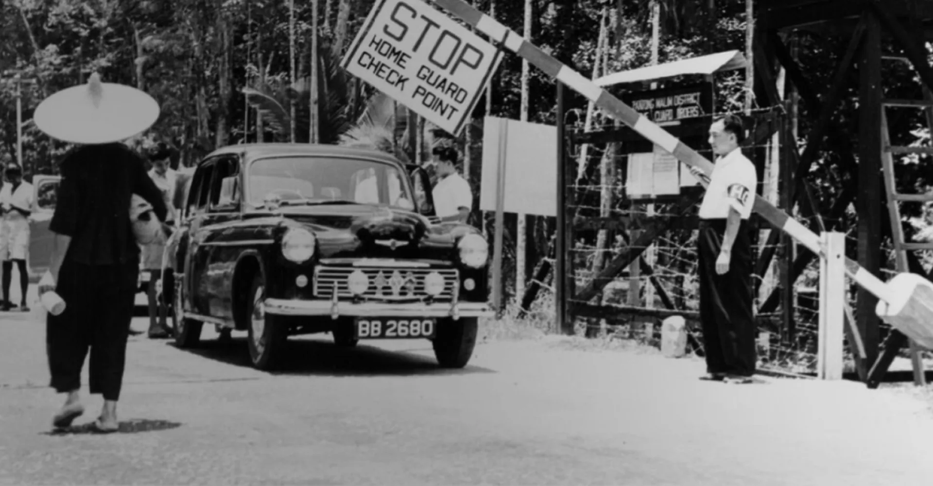 A vintage black and white photo depicts a checkpoint with a car, a stop sign, and a guard, surrounded by dense foliage.