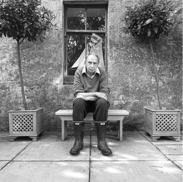 A person in gardening boots sits on a bench, surrounded by potted plants, in front of a rustic stone wall and window.