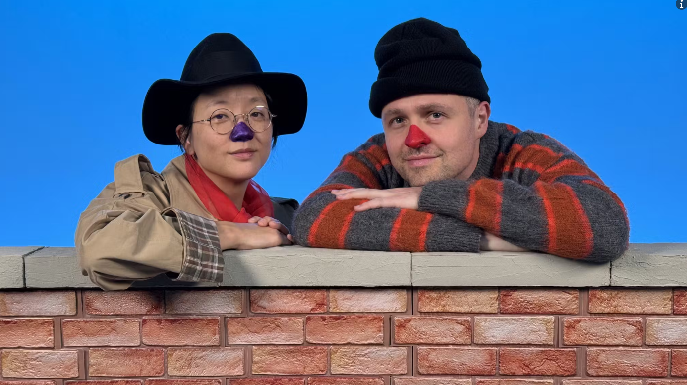 Two people in front of a bright blue background, resting their arms on a brick wall, one wears a cowboy hat and the other a beanie.