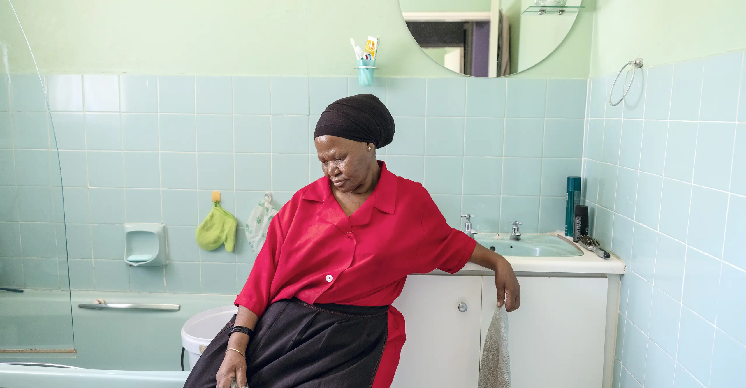 A woman in a red shirt and black skirt sits on the edge of a bathroom sink, surrounded by aqua tiles and bathroom essentials.