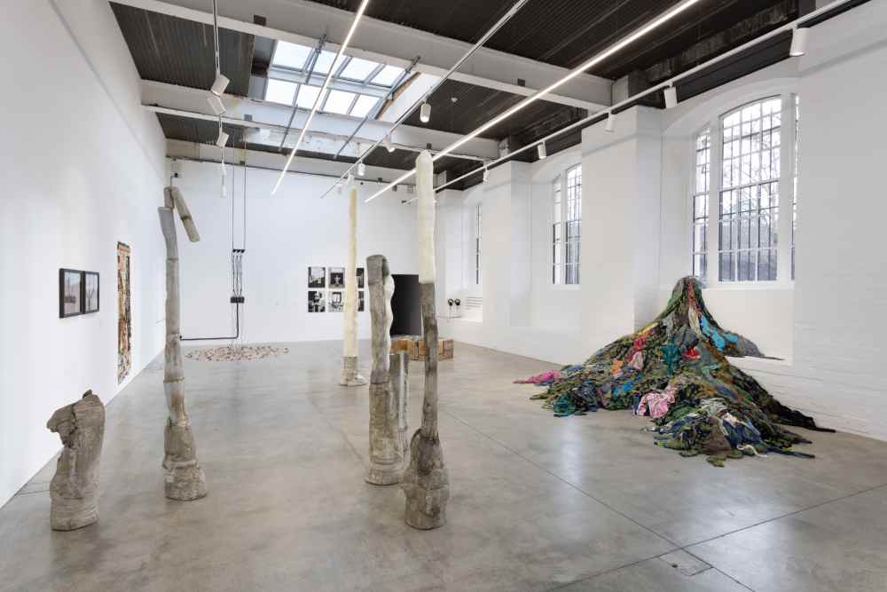 A modern art gallery featuring abstract sculptures, photographs on walls, and a colorful textile mound on the floor.