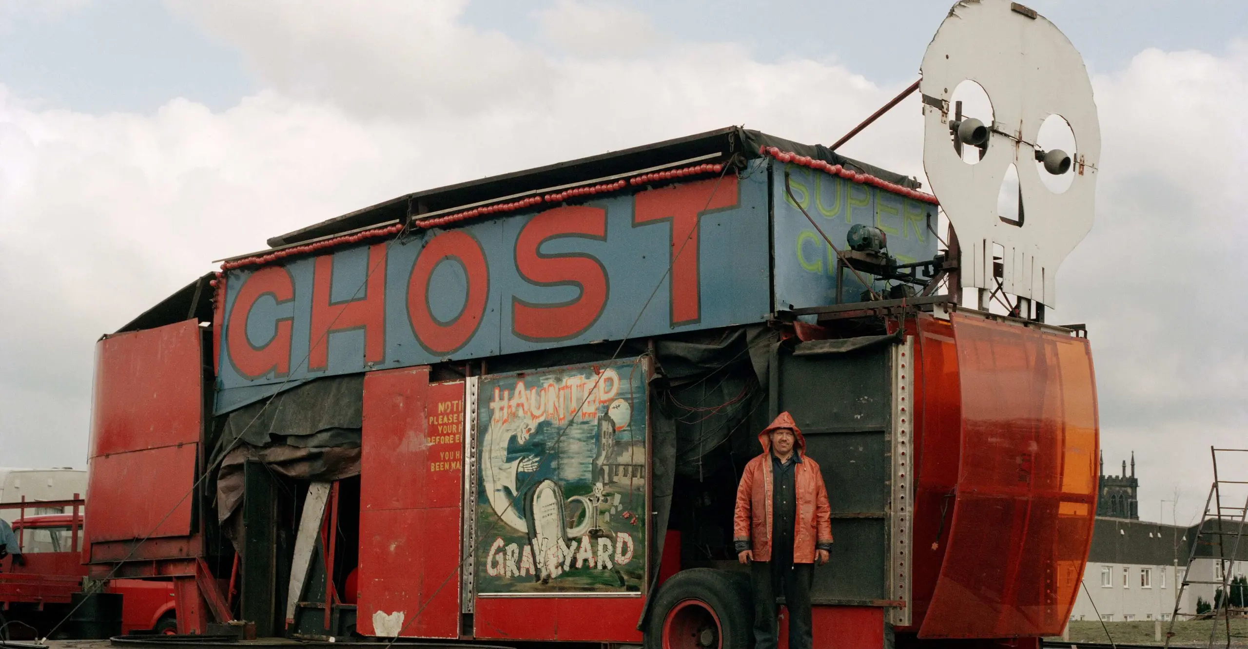 A vintage carnival attraction with a vibrant "GHOST" sign, featuring a skull decoration and eerie haunted graveyard artwork.
