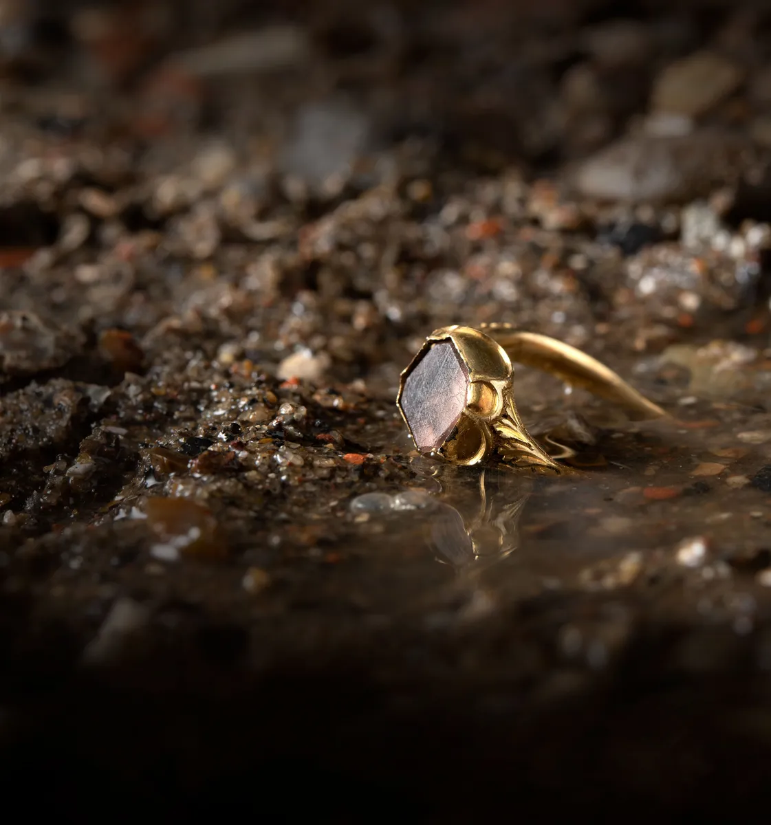 A close-up of a gold ring with a unique hexagonal stone, resting on a textured, gritty surface, reflecting in shallow water.