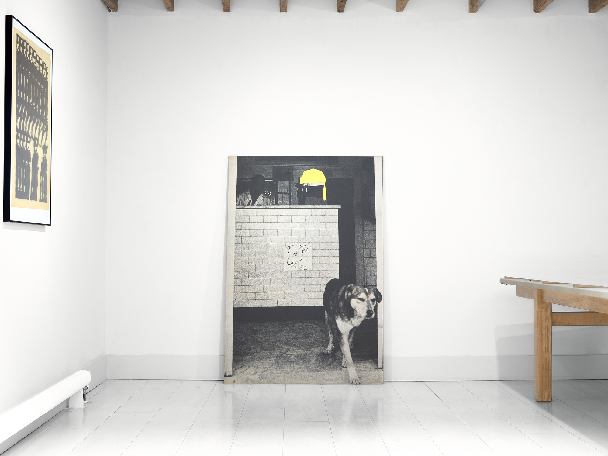 A dog stands in front of a large black-and-white artwork depicting a man and dog behind a wall, in a minimalist gallery space.