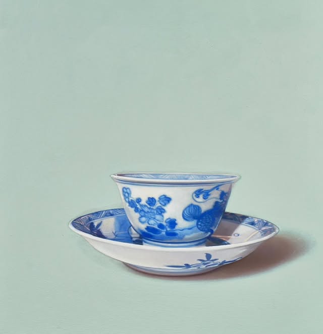 A delicate blue and white porcelain bowl sits atop a matching saucer, set against a soft green background.