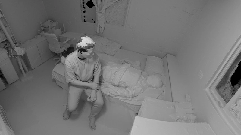 A monochromatic room with a seated person beside a wrapped figure on a bed, surrounded by scattered furniture and a window.