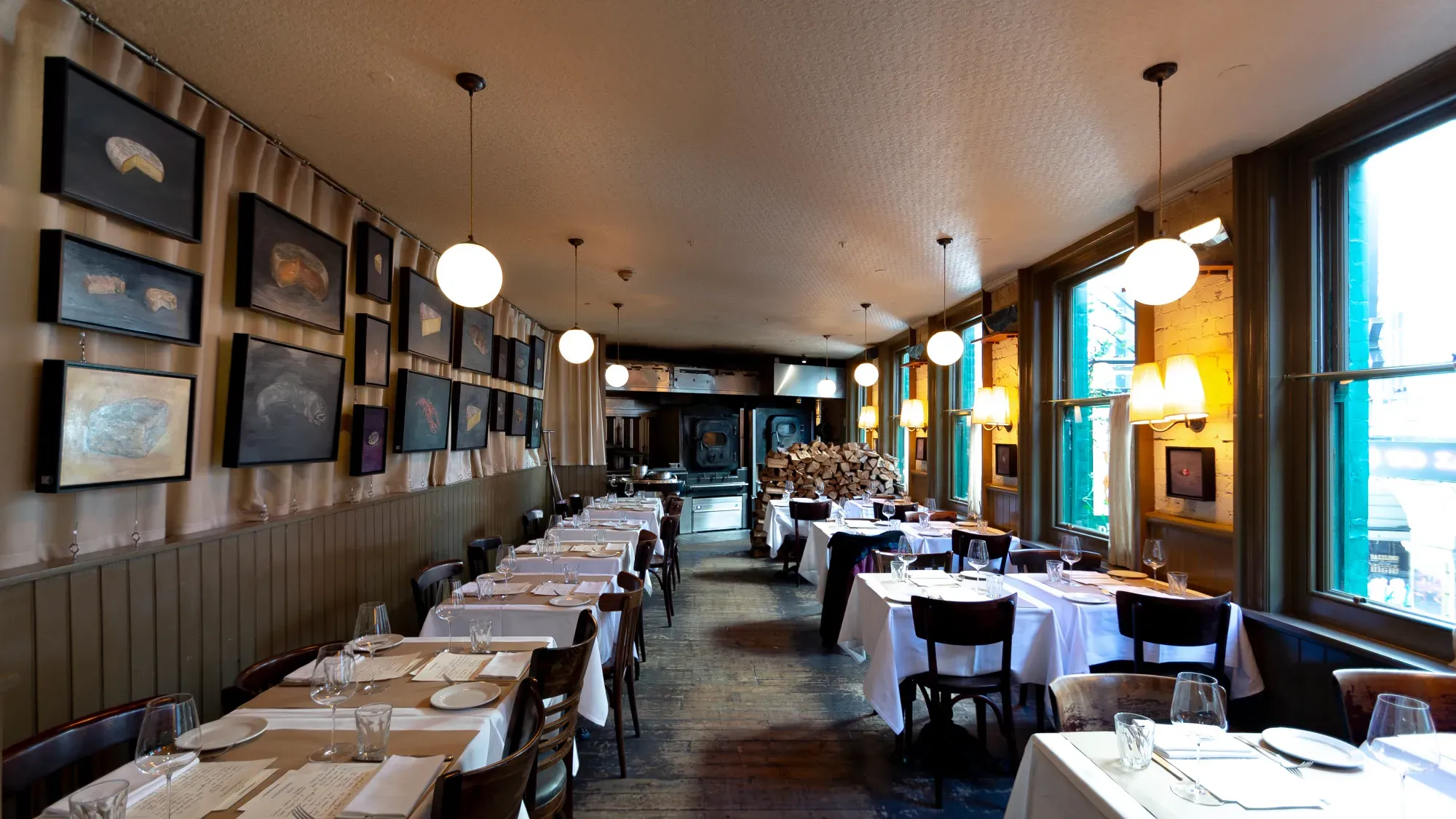 A cozy restaurant interior featuring white tablecloths, round pendant lights, and framed art on the walls, with a woodpile in the back.