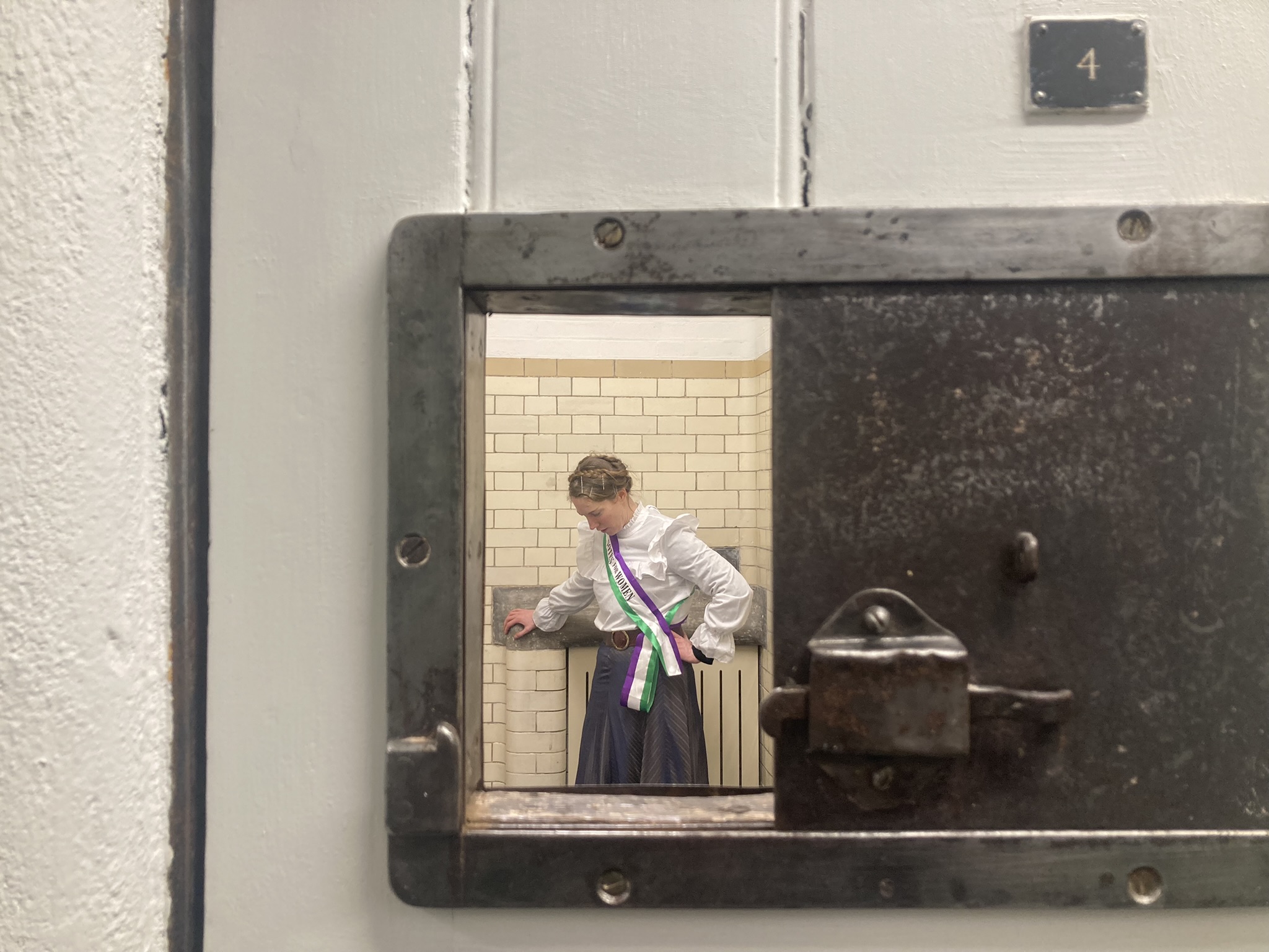A person in a historic jail cell, wearing a white top and a dark skirt, with a colorful sash, standing near a tiled wall.