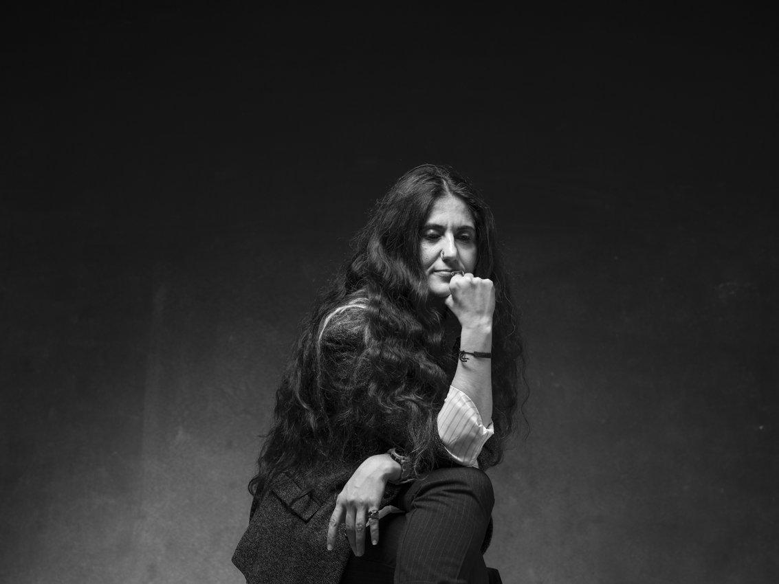 A person with long, curly hair is seated in a contemplative pose, dressed in a classic outfit against a dark background.