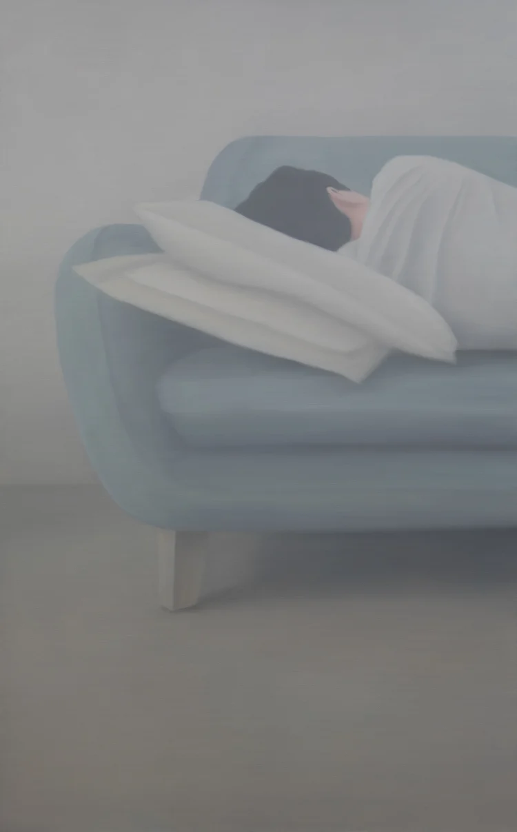 A person curled up on a light blue couch, wrapped in a white blanket, resting on multiple pillows against a soft, muted background.