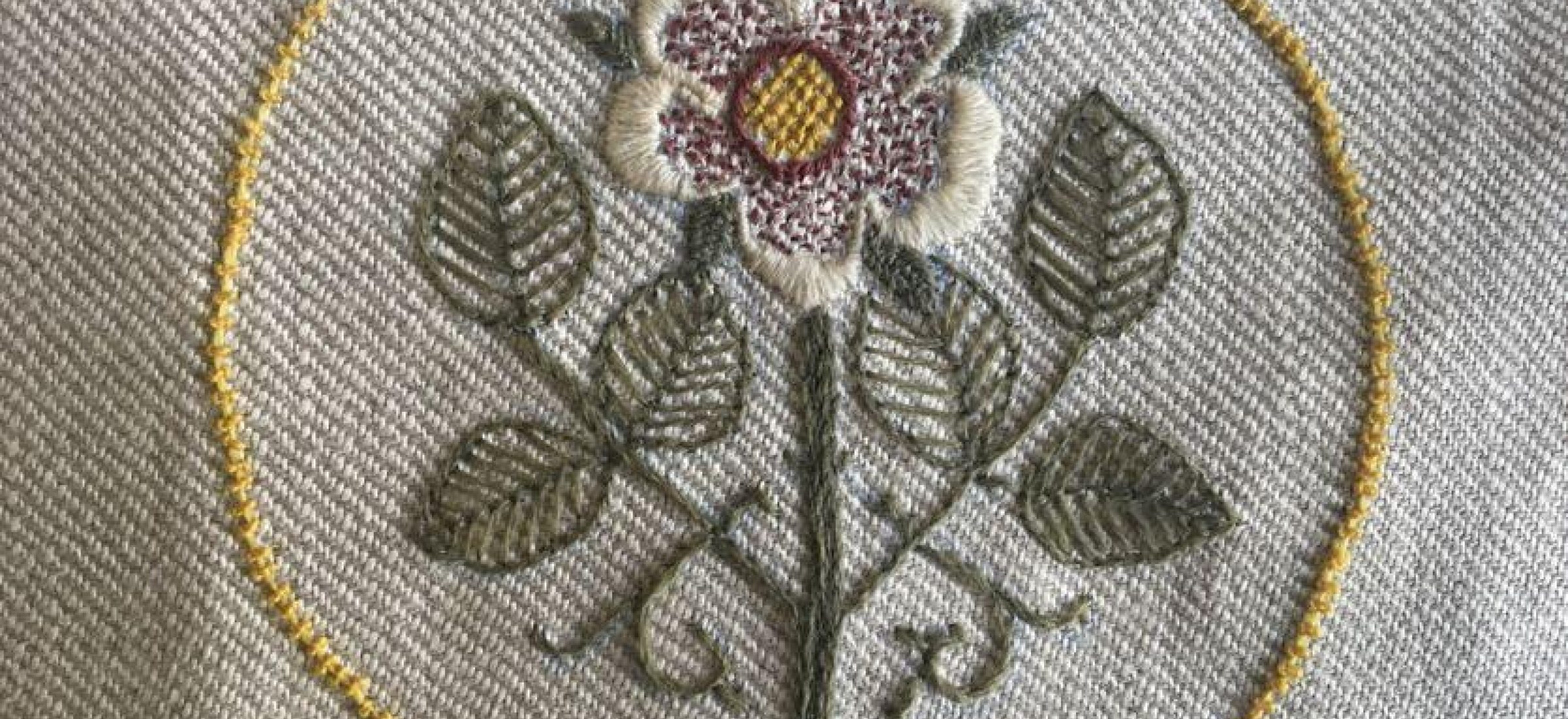 A detailed embroidery of a flower with leaves, surrounded by a yellow circular stitch, on a textured gray fabric background.