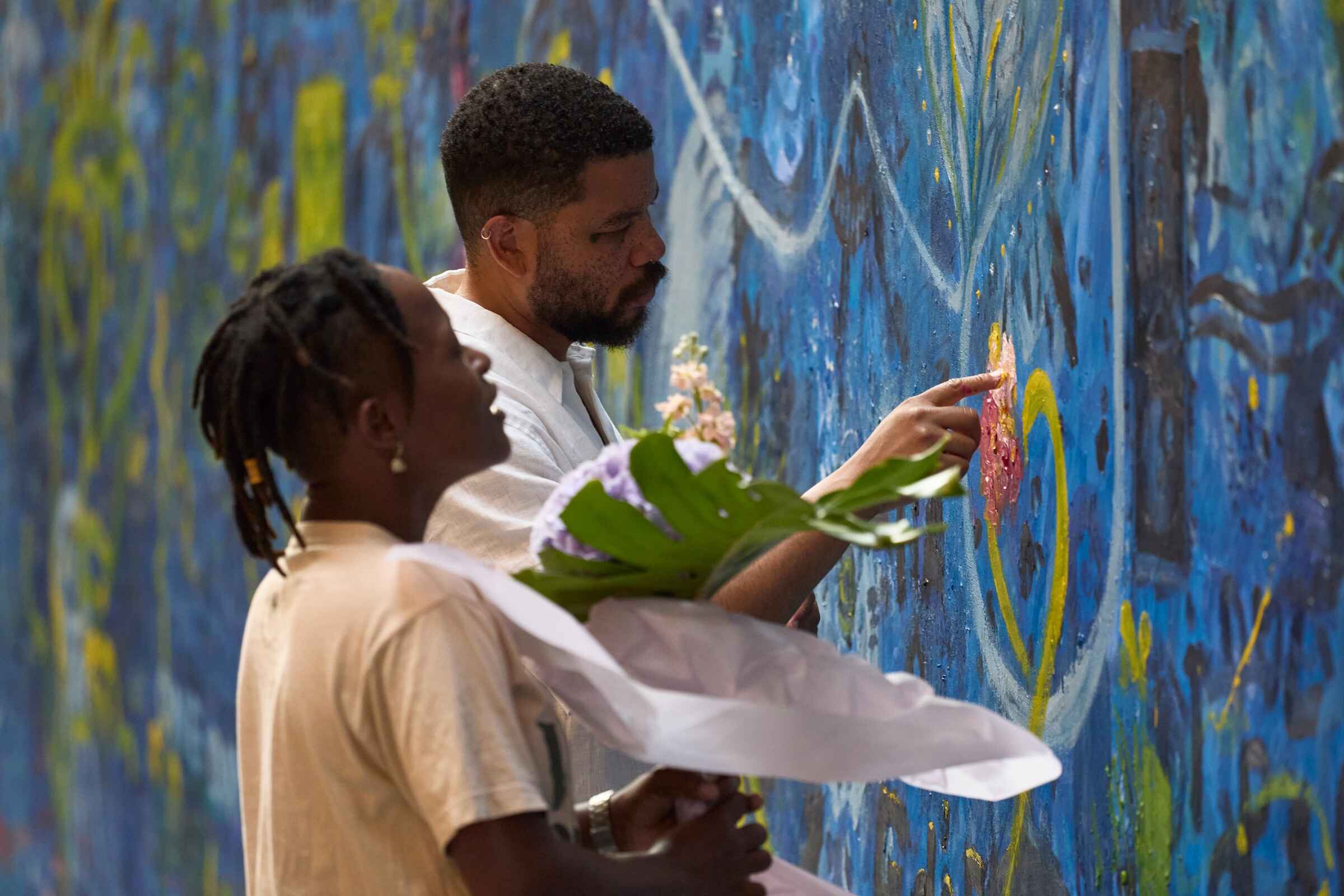 Two individuals admire colorful street art on a vibrant wall, one pointing at a design while holding a bouquet of flowers.