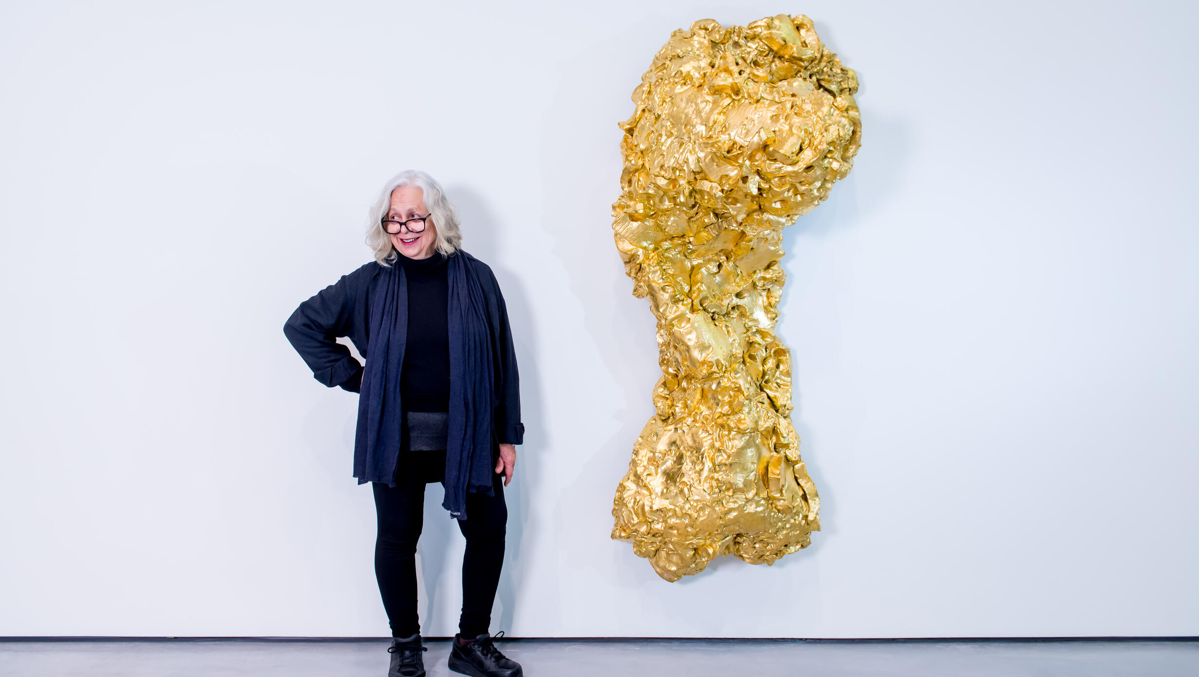 A woman stands beside a large gold sculpture, showcasing its intricate design and gleaming surface.