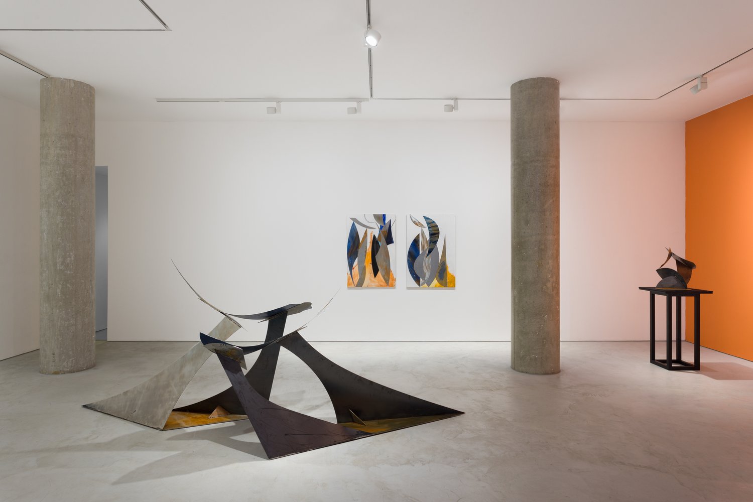 An art gallery featuring abstract sculptures and paintings on white walls, with a bold orange accent wall and exposed concrete columns.