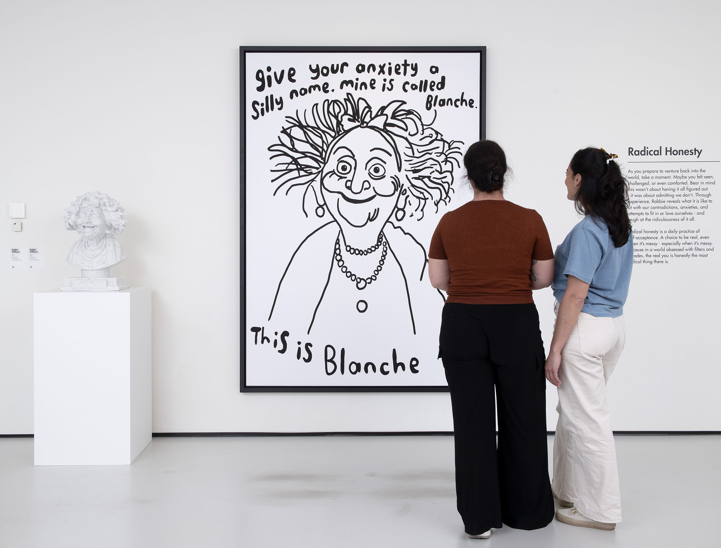 Two women stand before a large illustrated artwork with text about anxiety, titled "This is Blanche," in a bright gallery setting.