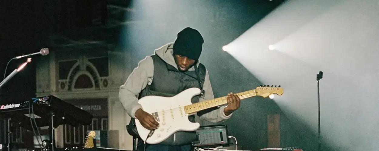 A musician plays an electric guitar on stage, illuminated by dramatic lighting, with a keyboard setup nearby.