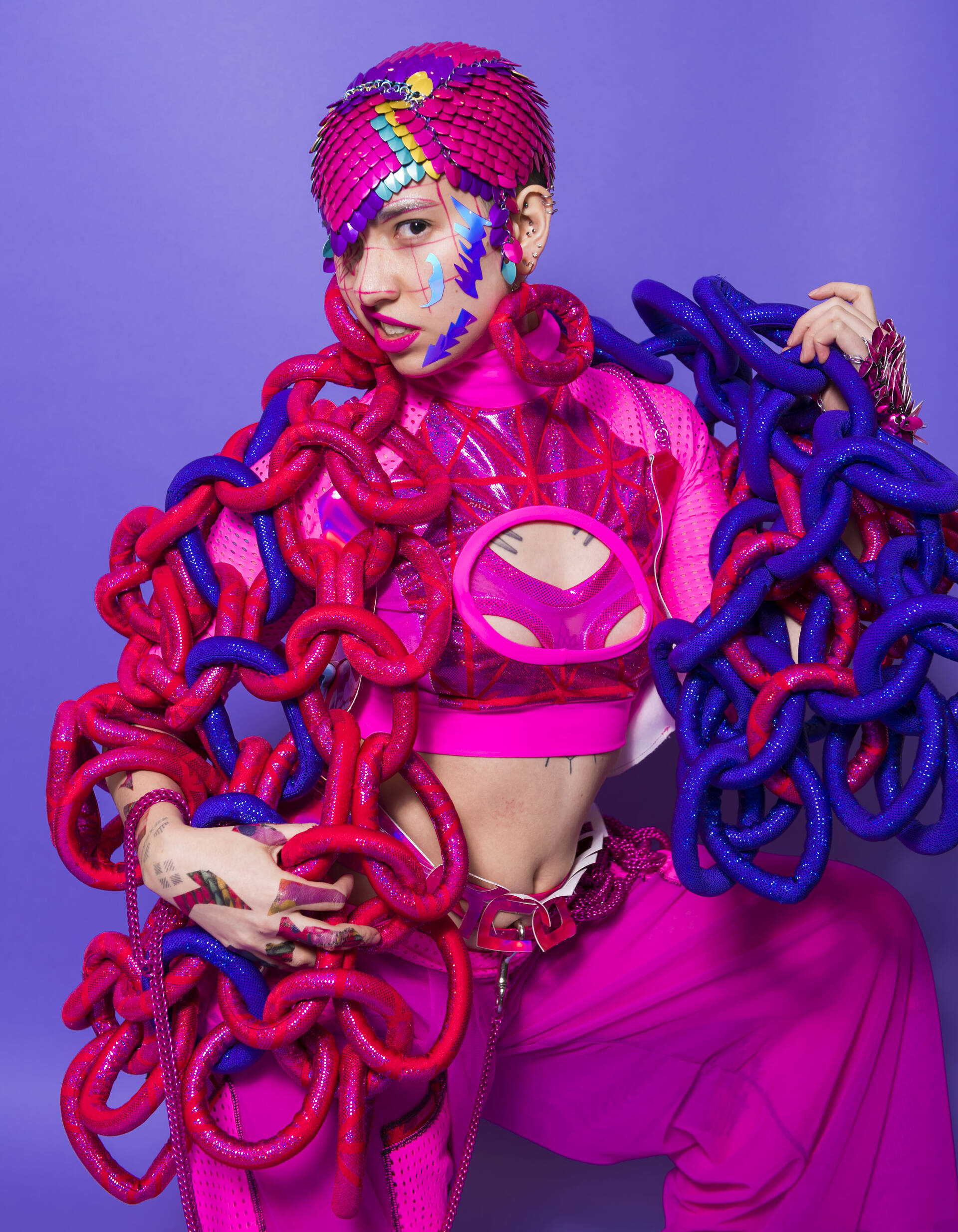 A fashion model adorned in vibrant pink and purple chains poses against a bold purple backdrop, showcasing a unique and artistic style.