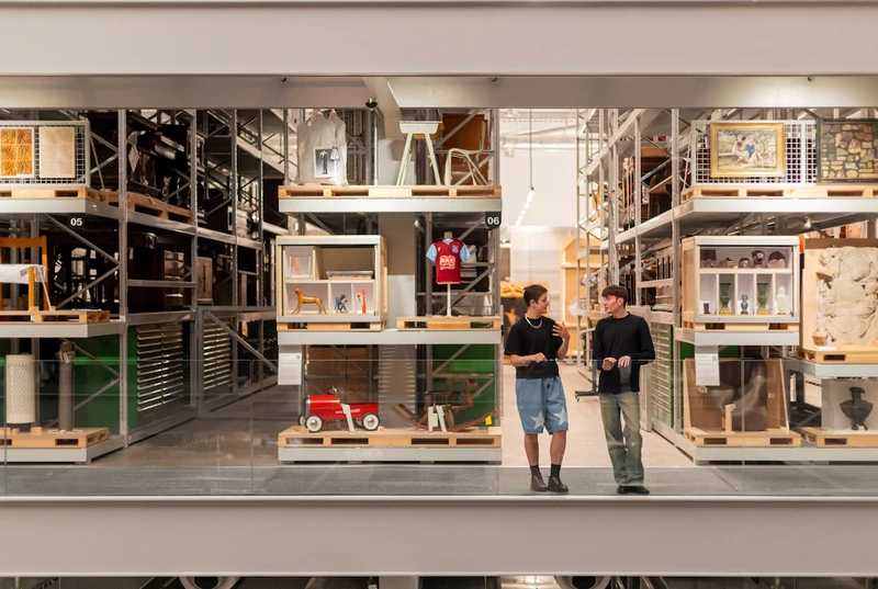 Two young adults converse in an exhibition space filled with organized shelves displaying a variety of artifacts and artworks.