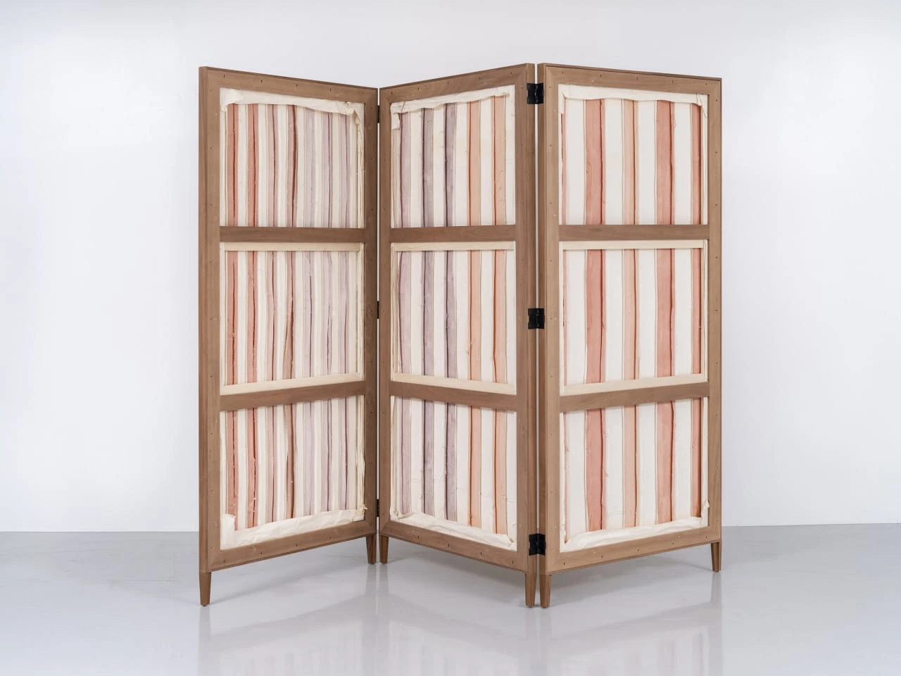 A three-panel folding screen with a wooden frame featuring alternating stripes of soft peach and cream colors on its fabric.