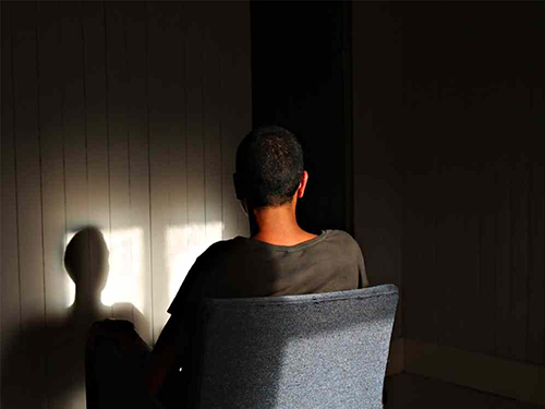 A person sits on a chair, viewed from behind, in a softly lit room casting shadows on the wall.