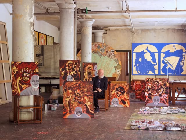An artist sits in a spacious studio surrounded by vibrant, abstract paintings and art supplies, with tall columns supporting the space.