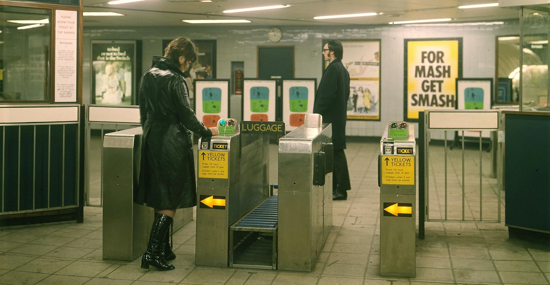 A woman in a black coat stands at a subway turnstile, while another man dressed in formal attire waits nearby, surrounded by vintage posters.