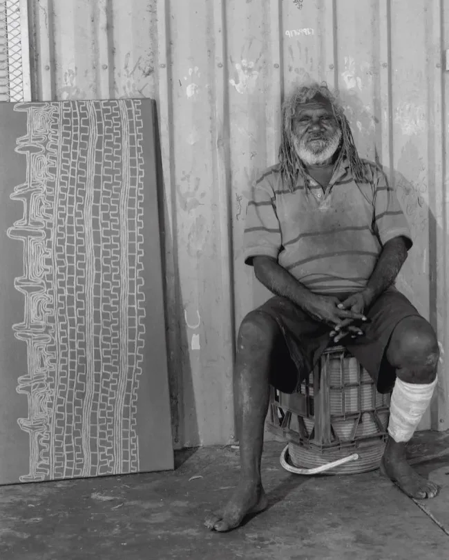 Elderly Indigenous man sits on a crate, wearing a striped shirt and a bandage on his leg, next to a large artwork depicting intricate tire-track patterns. | 2025 Unit