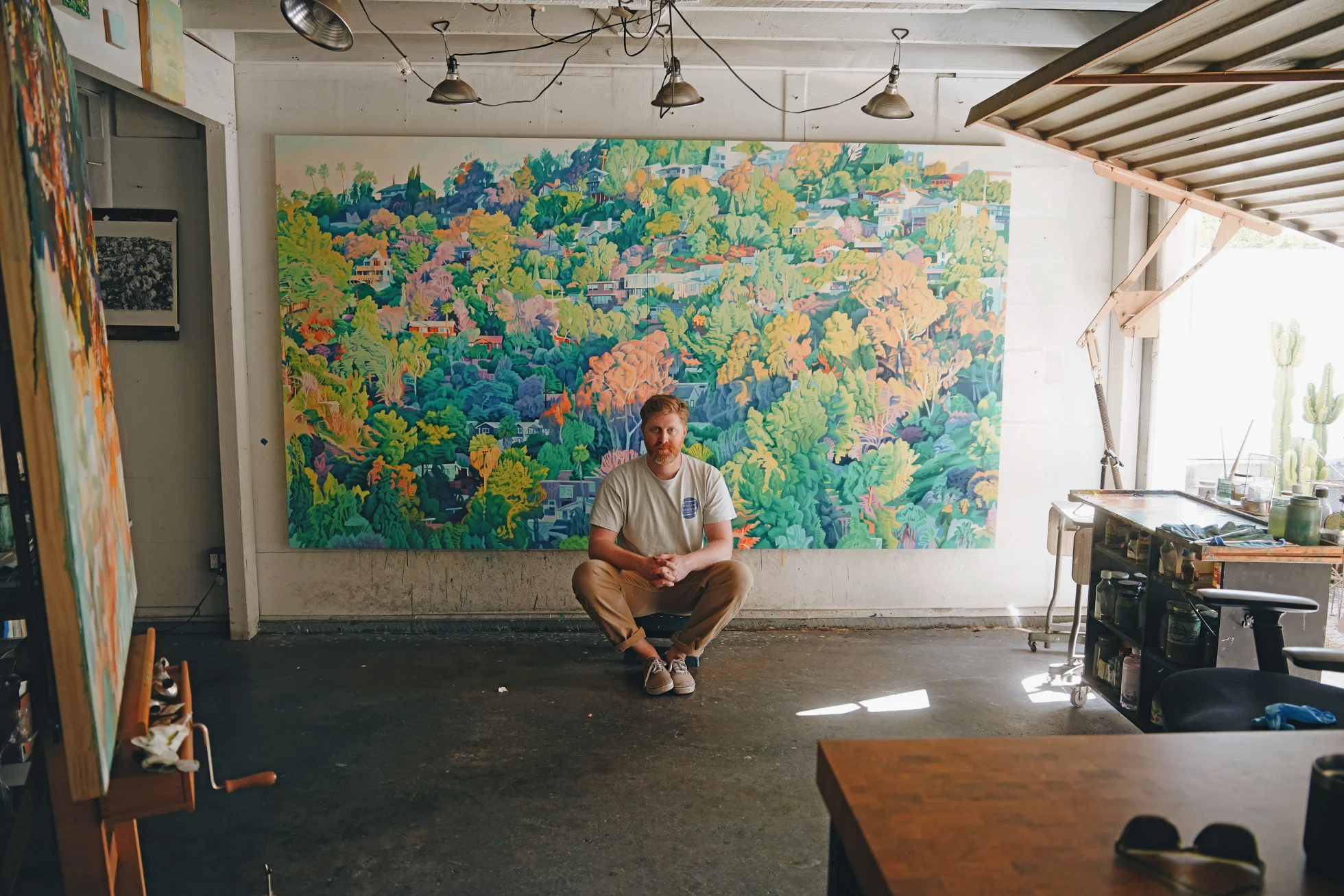 Man sits in an art studio, facing a large vibrant landscape painting of trees and houses.