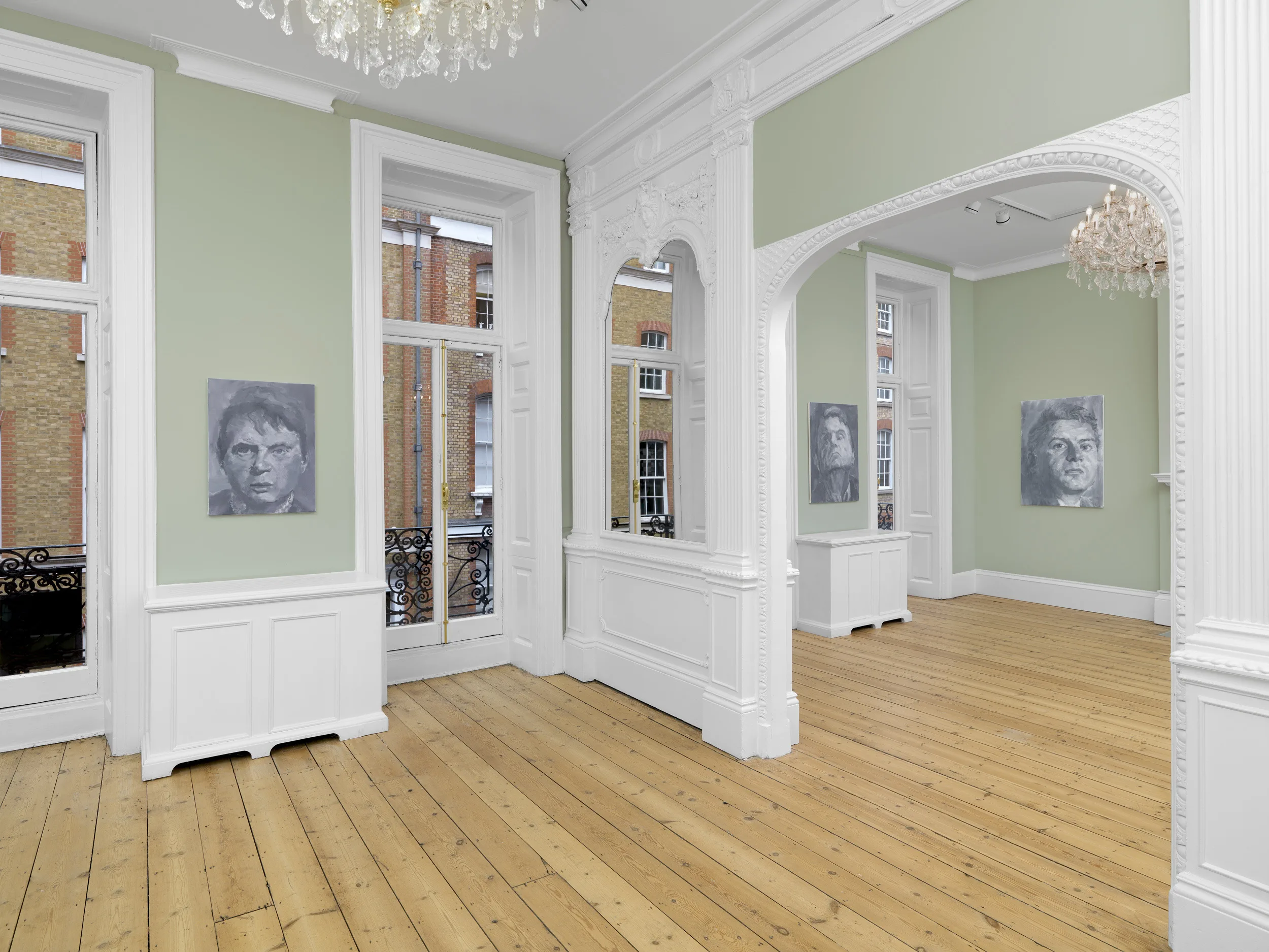 A light green gallery space featuring tall windows, polished wooden floors, decorative molding, and artwork on the walls.