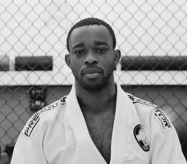 A person wearing a white Brazilian jiu-jitsu gi stands in front of a chain-link fence, exuding confidence and determination.