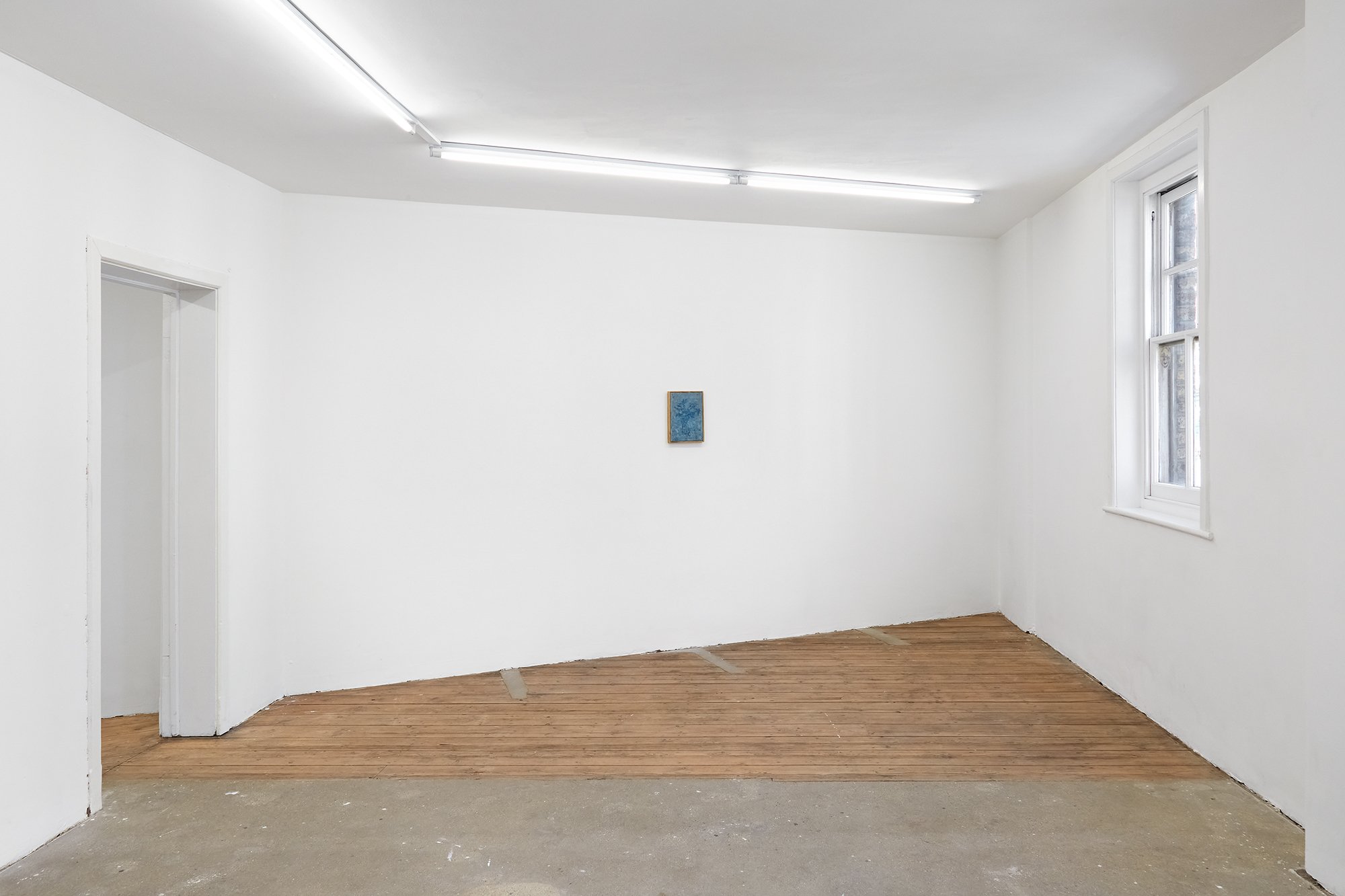 A minimalist art gallery featuring white walls, wooden flooring, a window, and a small blue artwork hung on the wall.