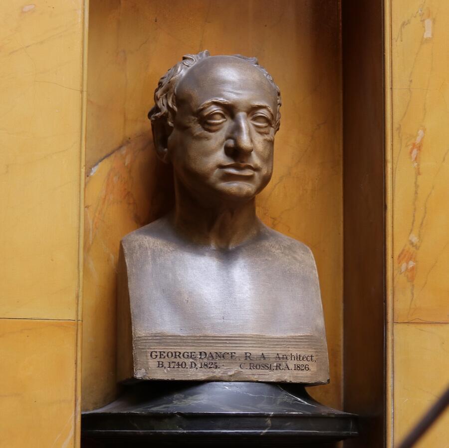 A bronze bust of George Dance, an architect, displayed on a dark pedestal against a vibrant yellow marble wall.