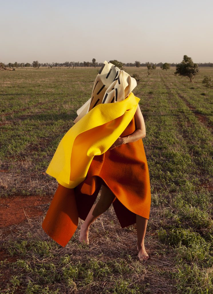 A figure draped in vibrant yellow and orange fabric stands barefoot in a sunlit, grassy field, blending art and nature.