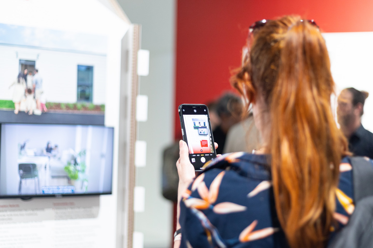 Person taking a picture of an interior design display on a smartphone, with a home design on a screen in the background.