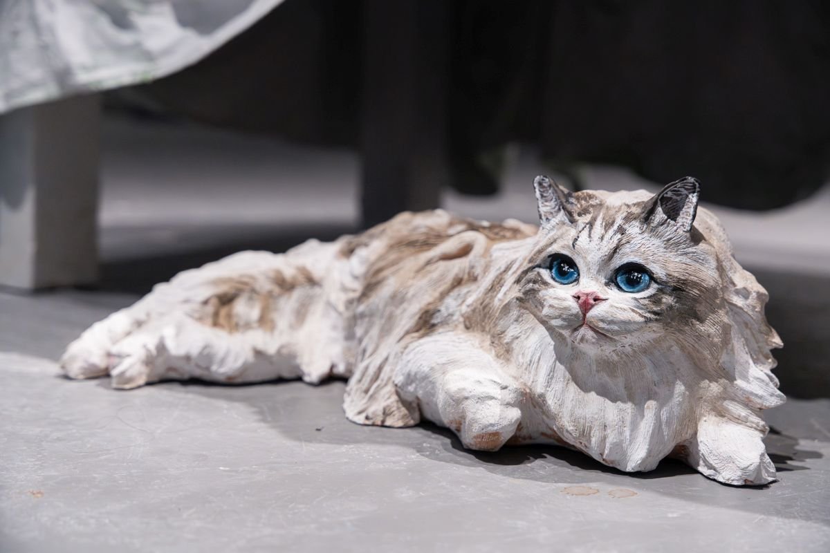 Sculptural cat figurine, light gray and beige, with large blue eyes, resting on a gray floor.