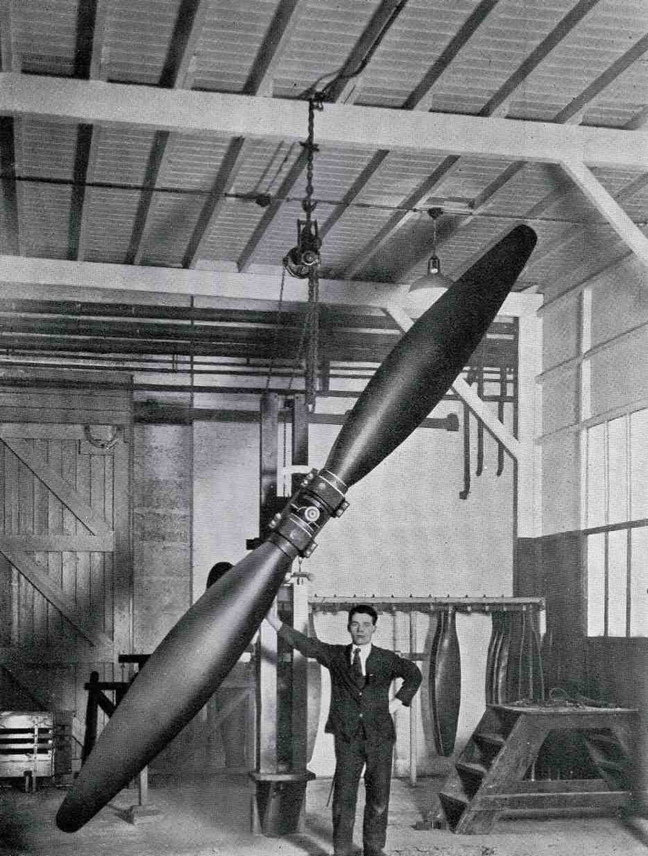 A man in a suit stands beside a large airplane propeller hanging from the ceiling in an industrial workshop, with machinery and equipment visible.