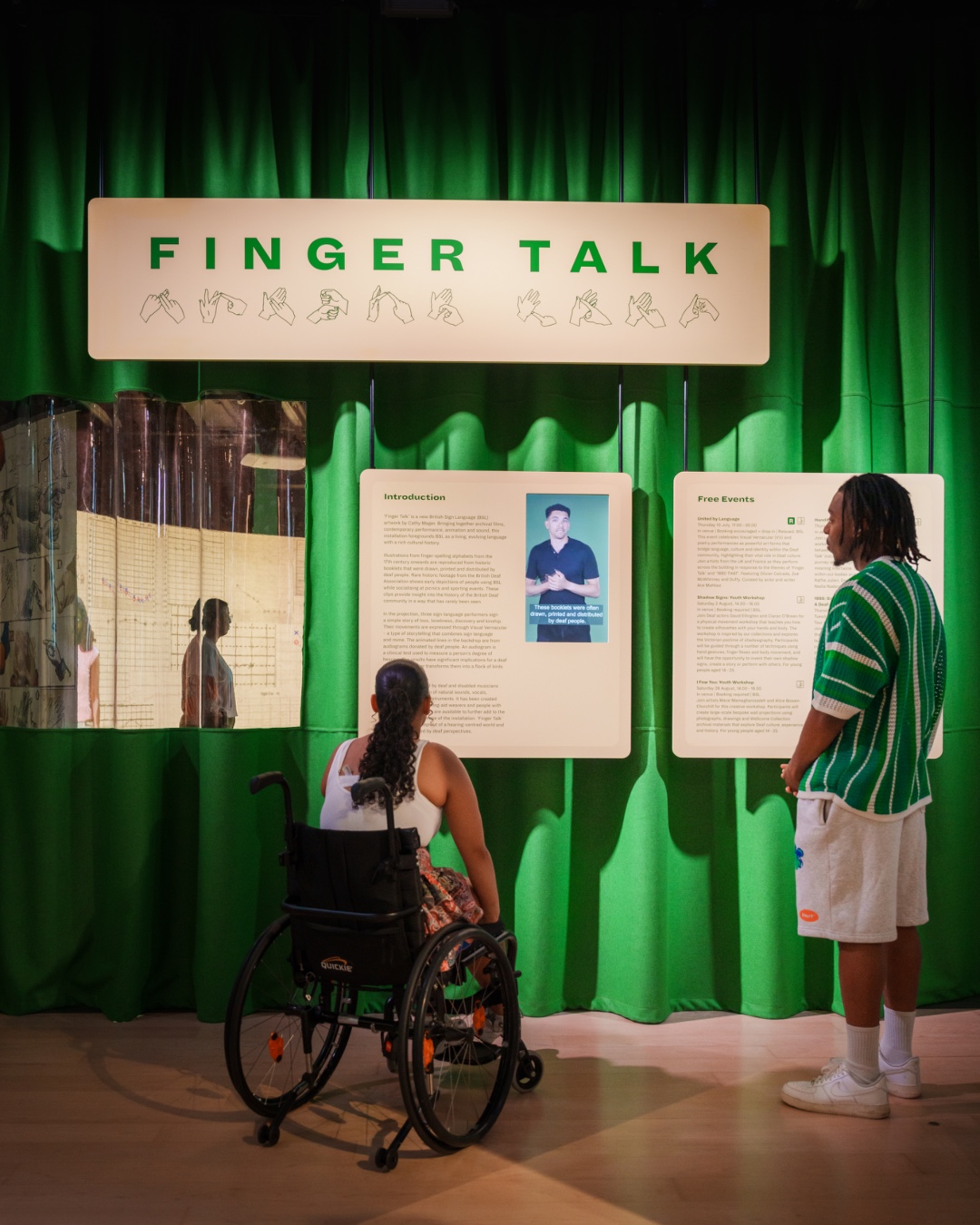 Two individuals, one in a wheelchair, view exhibits under a "FINGER TALK" sign against a green curtain backdrop.